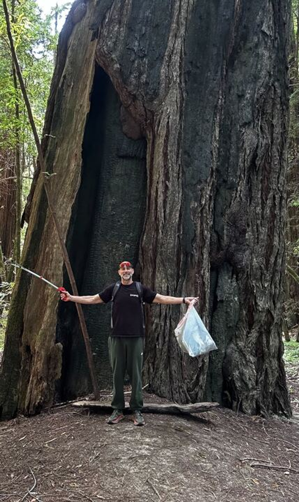 A man smiling for a photo in front of a large tree in a forest
