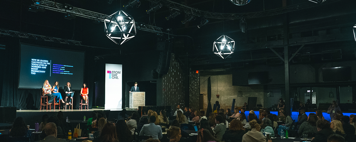 A panel of speakers discusses workforce and HR strategies on stage at a From Day One conference. The stage features a large screen displaying the session title and speaker names, while the audience listens attentively in a dimly lit venue with geometric light fixtures overhead.