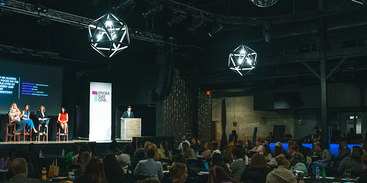 A panel of speakers discusses workforce and HR strategies on stage at a From Day One conference. The stage features a large screen displaying the session title and speaker names, while the audience listens attentively in a dimly lit venue with geometric light fixtures overhead.