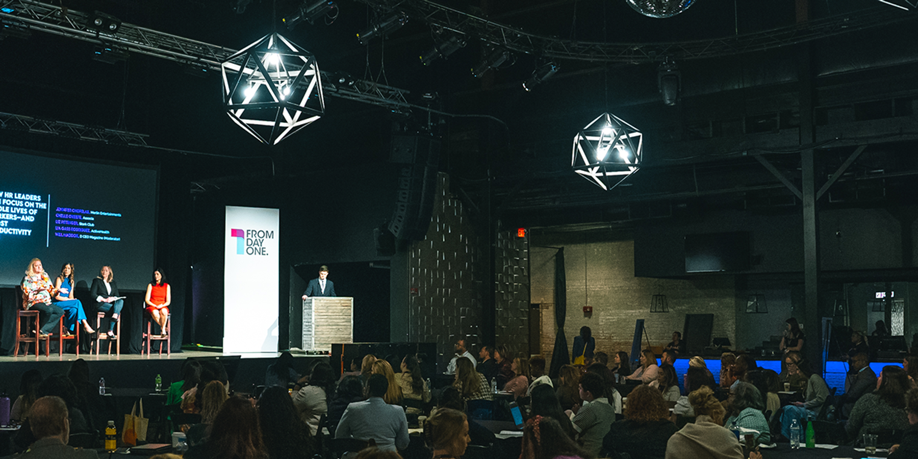 A panel of speakers discusses workforce and HR strategies on stage at a From Day One conference. The stage features a large screen displaying the session title and speaker names, while the audience listens attentively in a dimly lit venue with geometric light fixtures overhead.