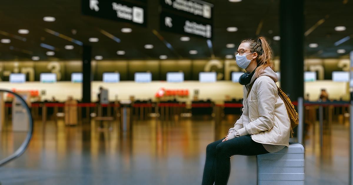 A person wearing a mask sitting on a suitcase at the airport
