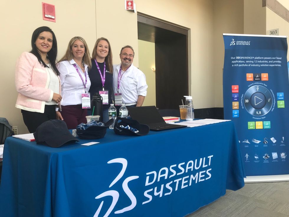 Four Dassault Syst\u00e8mes team members smiling for a picture behind their Dassault Syst\u00e8mes table at a conference 