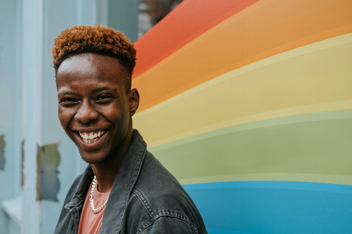 Man, smiling, with a PRIDE rainbow behind him