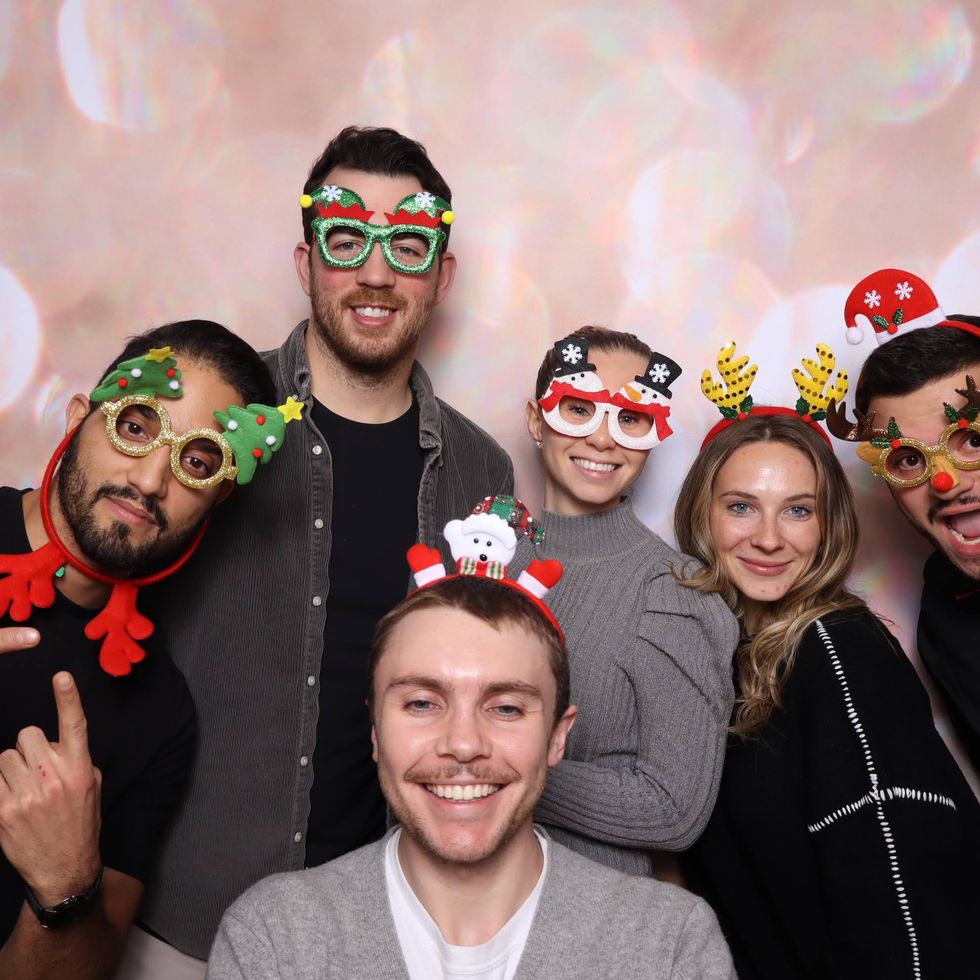 Six coworkers from Elastic posing together in festive headbands and holiday-themed glasses, smiling and leaning in for a playful group photo.