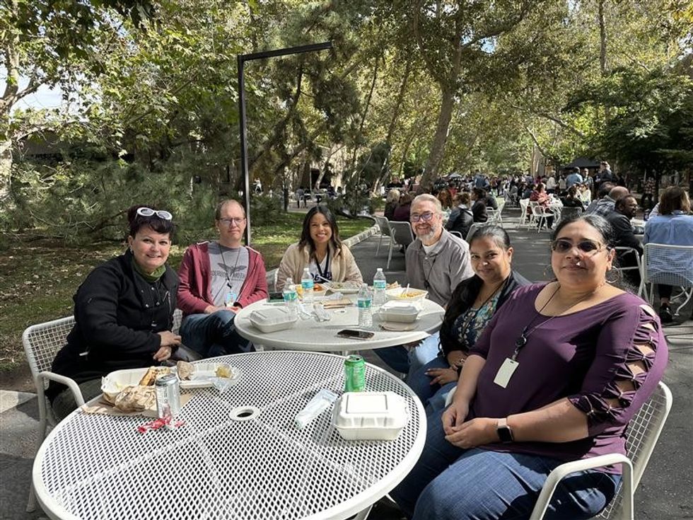 Six Esri employees smiling for a photo as they eat lunch together at an outside table in a park.