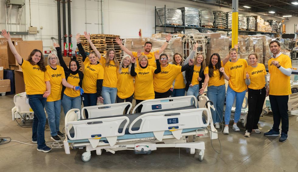 Stryker employees standing together in a warehouse facility with hospital beds, wearing yellow Stryker shirts and raising their hands, representing teamwork and collaboration across teams.